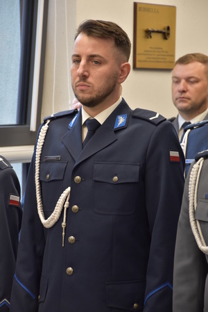 Zdjęcie kolorowe. Policjant w umundurowaniu wyjściowym koloru granatowego, ze sznurem. Obok niego i za nim stoją w rzędach policjanci uczestniczący w uroczystości.