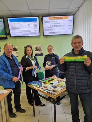 Zdjęcie kolorowe. Policjantka stojąca przy stoisku zorganizowanym na terenie Zakładu Ubezpieczeń Społecznych. Z prawej strony policjant stoi kobieta trzymająca ulotki oraz mężczyzna z trzymający w dłoni odblask. Z lewej strony stoi mężczyzna, trzymający w dłoniach elementy odblaskowe.