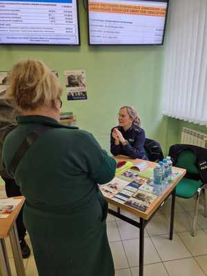 Zdjęcie kolorowe. Policjantka siedząca przy stoisku zorganizowanym na terenie Zakładu Ubezpieczeń Społecznych. Rozmawia z osobami stojącymi przy stoisku. Na stoisku ulotki, broszury i elementy odblaskowe przygotowane dla seniorów.