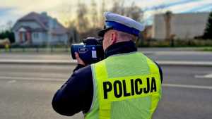 Zdjęcie kolorowe. Policjant mierzący prędkość pojazdów. Stojący tyłem. Ubrany w kamizelkę odblaskową koloru żółtego z napisem POLICJA