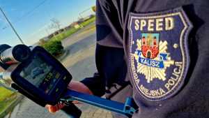 Zdjęcie kolorowe. Na pierwszym planie fragment rękawa munduru z naszywką na której znajduje się napis "Speed. Komenda Miejska Policji. Kalisz". Policjant trzyma w dłoni miernik prędkości, na którego wyświetlaczu znajduje się pojazd i pomiar prędkości -138 km/h