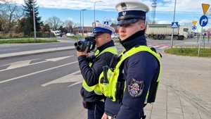 Zdjęcie kolorowe. Patrol dwuosobowy policjantów ruchu drogowego. Jeden z policjantów mierzy prędkość pojazdów poruszających się po drodze.