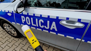 Zdjęcie kolorowe. Trzymane w dłoni urządzenie do badania stanu trzeźwości, na tle policyjnego oznakowanego radiowozu. Na drzwiach radiowozu napis POLICJA.
