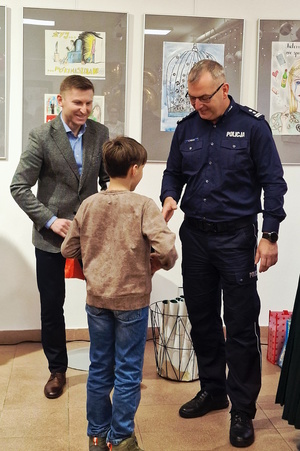 Zdjęcie kolorowe. Mały chłopiec odbiera nagrodę z rak mężczyzny. Stojący obok policjant gratuluje dziecku.
