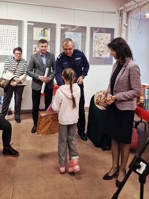 Zdjęcie kolorowe. Mała dziewczynka odbiera nagrodę z rąk mężczyzny. Stojący obok policjant gratuluje dziecku. Obok stoi kobieta, która przygląda się wręczeniunagrody.