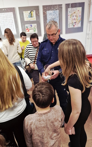 Zdjęcie kolorowe. Policjant trzyma w dłoniach przeźroczyste naczynie z losami. Dzieci zgromadzone wokół wyciągają z pojemnika losy.