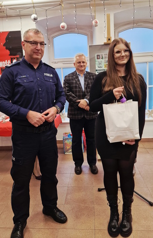 Zdjęcie kolorowe. Policjant i nastolatka pozują do zdjęcia po wręczeniu nagrody, którą w dłoniach trzyma dziewczyna. W tle pomiędzy pozującymi stoi mężczyzna.