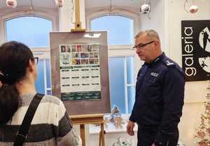 Zdjęcie kolorowe. Policjant rozmawia z kobietą stojąc przed sztalugą z kalendarzem na rok 2026, który zawiera prace laureatów konkursu "Wolność bez nałogów".