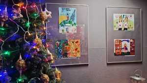 Zdjęcie kolorowe. Z lewej strony fragment choinki. Na ścianie w antyramach wiszą kolorowe prace laureatów konkursu wolność bez nałogów.