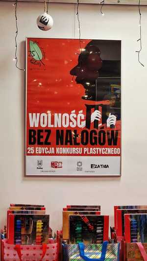 Zdjęcie kolorowe. Plakat konkursu plastycznego "Wolność bez nałogów"wiszący na ścianie. Pod spodem nagrody dla laureatów.