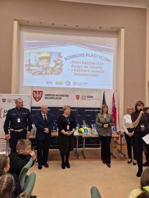 Zdjęcie kolorowe. Kobiety i mężczyźni, przedstawiciele instytucji organizujący konkurs "Moja bezpieczna droga do szkoły - w kasku zawsze bezpieczniej". Osoby stoją pod planszą z nazwą konkursu. Jedna z osób, kobieta mówi do mikrofonu trzymanego w dłoni.