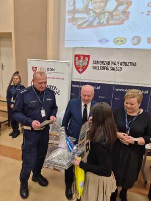 Zdjęcie kolorowe. Troje organizatorów konkursu - Moja bezpieczna droga do szkoły - w kasku zawsze bezpieczniej, wręcza nagrody uczestniczce konkursu.