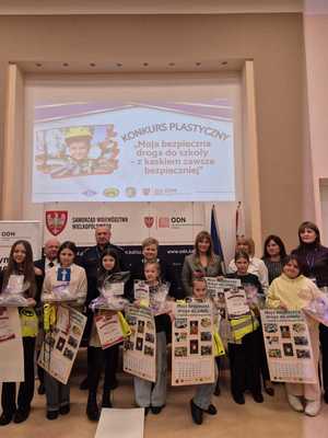 Zdjęcie kolorowe. Organizatorzy konkursu - Moja bezpieczna droga do szkoły - w kasku zawsze bezpieczniej, wspólnie z nagrodzonymi uczestnikami pozują do zdjęcia. Dzieci trzymają w dłoniach nagrody i dyplomy.