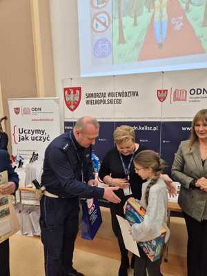 Zdjęcie kolorowe. Troje organizatorów konkursu - Moja bezpieczna droga do szkoły - w kasku zawsze bezpieczniej, wręcza nagrody uczestniczce konkursu.