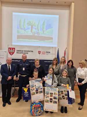 Zdjęcie kolorowe. Organizatorzy konkursu - Moja bezpieczna droga do szkoły - w kasku zawsze bezpieczniej, wspólnie z nagrodzonymi uczestnikami pozują do zdjęcia. Dzieci trzymają w dłoniach nagrody i dyplomy.