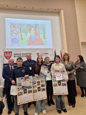 Zdjęcie kolorowe. Organizatorzy konkursu - Moja bezpieczna droga do szkoły - w kasku zawsze bezpieczniej, wspólnie z nagrodzonymi uczestnikami pozują do zdjęcia. Dzieci trzymają w dłoniach nagrody i dyplomy.