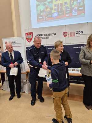 Zdjęcie kolorowe. Troje organizatorów konkursu - Moja bezpieczna droga do szkoły - w kasku zawsze bezpieczniej, wręcza nagrody uczestnikowi konkursu.