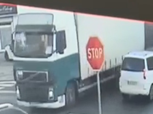 Zdjęcie kolorowe. Pojazd ciężarowy stojący przodem. Obok pojazdu stoi samochód osobowy. Pomiędzy pojazdami znak STOP.