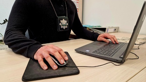 Zdjęcie kolorowe. Osoba siedząca przy biurku, przed komputerem. Widać jedynie tors osoby i ręce. Na szyi zawieszone etui z policyjną "blachą"