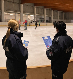 Zdjęcie kolorowe. Policjantki odwrócone tyłem na tle lodowiska, na którym jeżdżą osoby. W dłoniach trzymają plakaty i ulotki.