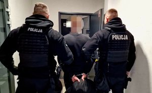 Zdjęcie kolorowe. Dwaj umundurowani trzymają zatrzymanego, który stoi pomiędzy nimi. Policjanci wraz zatrzymanym wchodzą do pomieszczeń dla osób zatrzymanych. Zatrzymany ma założone kajdanki na ręce trzymane z tyłu.