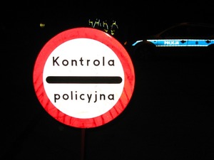 Zdjęcie kolorowe. Na pierwszym planie znak drogowy z napisem kontrola policyjna. W tle bok oznakowanego radiowozu z napisem Policja na drzwiach.