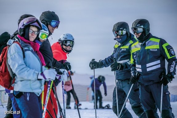 Zdjęcie kolorowe. Dwu osobowy, narciarski patrol policjantów na stoku. Funkcjonariusze w umundurowaniu służbowym z kaskami na głowach. Policjanci pozują do fotografii z pięcioosobową grupą narciarzy.