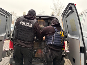Zdjęcie kolorowe. Dwaj policjanci zamykają tylne drzwi od przedziału w radiowozie, gdzie znajduje się osoba zatrzymana.