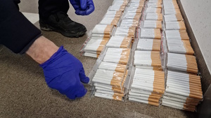 Zdjęcie kolorowe, na którym widoczne są dłonie w rękawiczkach jednorazowych koloru niebieskiego układające na podłodze woreczki foliowe z papierosami.