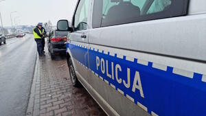Zdjęcie kolorowe. Na pierwszym planie z prawej strony policyjny, oznakowany radiowóz z napisem POLICJA. Przed radiowozem, zaparkowany samochód osobowy obok którego stoi dwójka policjantów. Z lewej strony droga po której poruszają się pojazdy.