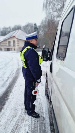 Zdjęcie kolorowe. Policjant z Wydziału Ruchu Drogowego rozmawia z kierowcą busa, który siedzi za kierownicą pojazdu. Policjant w lewej dłoni trzyma tarczę służącą do zatrzymywania pojazdów.