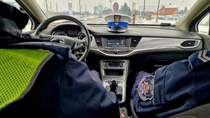 Zdjęcie kolorowe. Wnętrze radiowozu. Z przodu, za kierownicą i na miejscu pasażera siedzą policjanci, którym widać tylko ramiona. Z przodu na podszybiu umieszczona jest policyjna czapka.