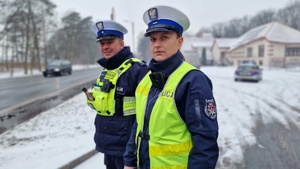 Zdjęcie kolorowe. Policjantka i policjant stoją na poboczu drogi i obserwują jezdnię, po której poruszają się pojazdy. Policjantka na lewym ramieniu  ma naszywkę z napisem Kalisz Komenda Miejska Policji.