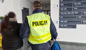 Zdjęcie kolorowe. Policjant prowadzi z lewej strony zatrzymaną kobietę. Z prawej strony na ścianie widoczna jest część tablicy informacyjnej z numerami pokoi i nazwami wydziałów