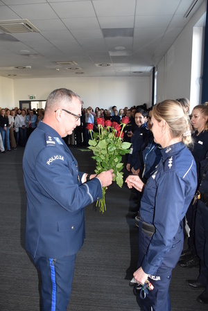Zdjęcie kolorowe. Policjant trzyma w dłoni bukiet czerwonych róż i wręcza je Paniom obecnym na sali.