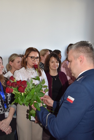 Zdjęcie kolorowe. Policjant trzyma w dłoni bukiet czerwonych róż i wręcza je Paniom obecnym na sali.