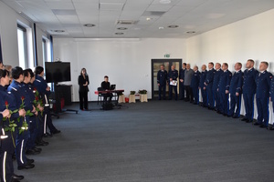 Zdjęcie kolorowe. Sala. Po prawej i lewej stronie stoją funkcjonariusze Policji. W środkowej części duet - kobieta (śpiewa) i mężczyzna (instrument klawiszowy)