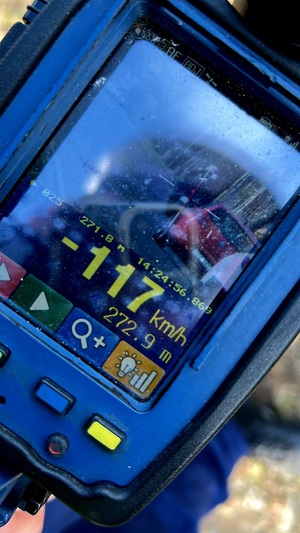 Zdjęcie kolorowe. Wyświetlacz ręcznego miernika prędkości, na którym widnieje zdjęcie motocyklisty i jadących za nim dwóch pojazdów. W dolnej części pomiar prędkości - 117 km/h. Nad nim godzina pomiaru 14:24:56,869, pod nim odległość z której mierzono pojazd 272.9 m