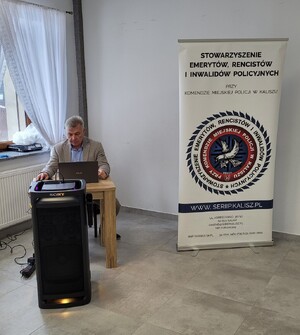 Zdjęcie kolorowe. Mężczyzna siedzący przy stole na którym stoi laptop. Przed stołem znajduje się duży głośnik z prawej strony baner Stowarzyszenia Emerytów, Rencistów i Inwalidów Policyjnych przy Komendzie Miejskiej Policji w Kaliszu.