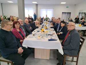 Zdjęcie kolorowe. Uczestnicy spotkania siedzą przy stołach na których przygotowane są przekąski i napoje.