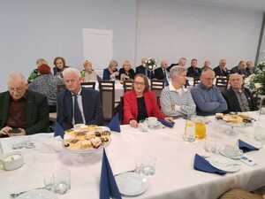 Zdjęcie kolorowe. Uczestnicy spotkania siedzą przy stołach na których przygotowane są przekąski i napoje.