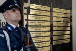 Zdjęcie kolorowe. Na pierwszym planie policjant w umundurowaniu wyjściowym z czapką w tle ściana, na której przymocowane są tabliczki wielkopolskich policjantów poległych na służbie.