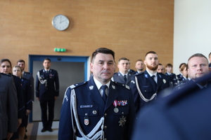 Zdjęcie kolorowe. w umundurowaniu wyjściowym ze sznurem przyjmuję meldunek od dowódcy uroczystości. W tle stoją awansowani policjanci.