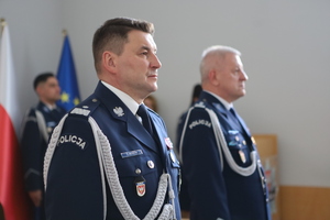 djęcie kolorowe. Komendant i przewodniczący związków stoją obok siebie. Obaj ubrani w mundur wyjściowy ze sznurem. W tle widać rozmazaną postać oraz flagi Unii Europejskiej oraz Polski.