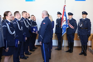 Zdjęcie kolorowe Komendant i przewodniczący związków wręczają policjantom teczki z aktami mianowania. W tle stoi poczet sztandarowy.