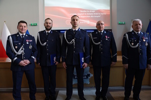 Zdjęcie kolorowe. Pięciu policjantów ubranych w mundury wyjściowe ze sznurem pozuje do zdjęcia. Dwóch z nich trzyma teczki z aktami mianowania. U góry na ekranie wyświetla się flaga Polski, na której w dolnej części jest napis: Służba to droga a każdy stopień to kolejny krok ku temu, by być wsparciem dla innych.