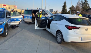 Zdjęcie kolorowe. Zdjęcie białego pojazdu i dwóch radiowozów oznakowanych. Obok białego pojazdu od strony pasażera stoją policjanci.