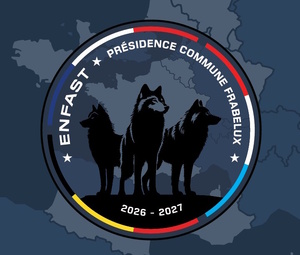 Grafika. Ciemne tło na którym narysowany są dwa okręgi. Zewnętrzny cały czarny, wewnętrzny przerywanymi kolorowymi liniami. W środku szkic trzech wilków. na około napisy  : ENFAST Presidence Commune Frabelux. 2026-2027
