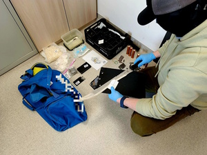 Zdjęcie kolorowe. Nieumundurowany policjant kuca nad rozłożonymi na podłodze przeźroczystymi woreczkami strunowymi z zawartością białej substancji. Policjant trzyma w dłoni maczetę zabezpieczoną podczas przeszukania. W prawym górnym roku skrzynka w której znajdują się kiełkujące rośliny marihuany. Z lewej niebieska, sportowa torba.