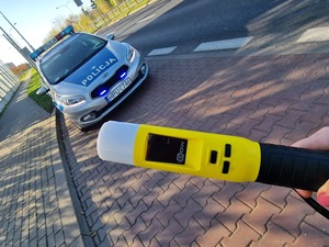 Zdjęcie kolorowe. Na pierwszym planie dłoń trzymająca urządzenie do badania stanu trzeźwości koloru żółtego. Na drugim planie zaparkowany, oznakowany policyjny radiowóz.